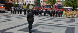 Na zdjęciu widać strażaków stojących na placu podczas uroczystości. Na pierwszym planie widać dowódcę uroczystości. Na drugim planie widoczni są strażacy ustawieni w pododdziale. Na kolejnym planie widoczne są pojazdy pożarnicze.