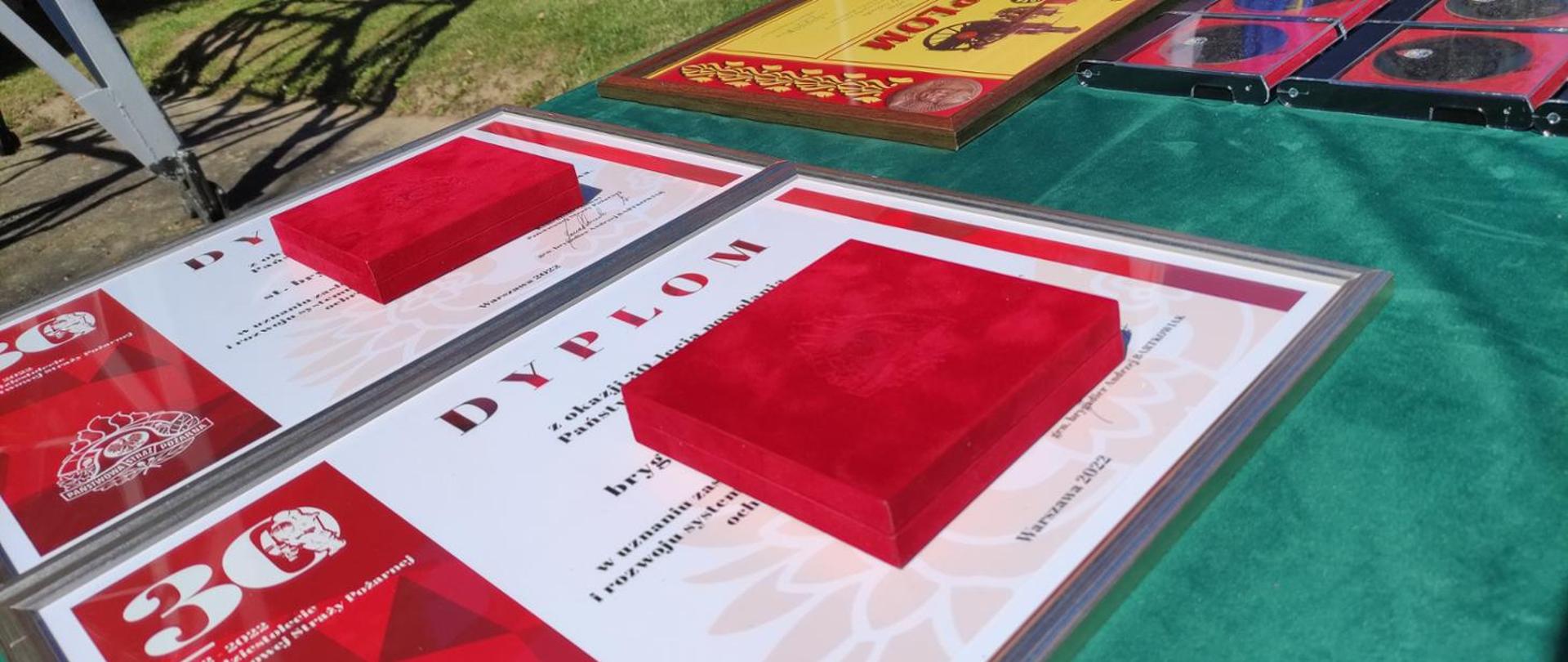 Na stole położone dyplomy z okazji 30-lecia powołania Państwowej Straży Pożarnej. Na dyplomach leżą zamknięte etui z medalami.