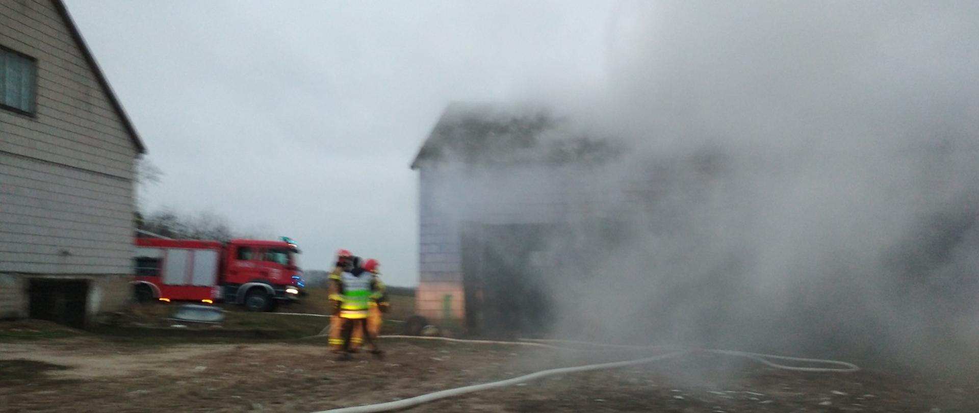 Na zdjęciu widać trzech strażaków, rozwinięty wąż strażacki, budynek murowanej stodoły z której wydobywa się szary dym, w lewej części zdjęcia część budynek mieszkalnego o białej ścianie, w tle samochód strażacki