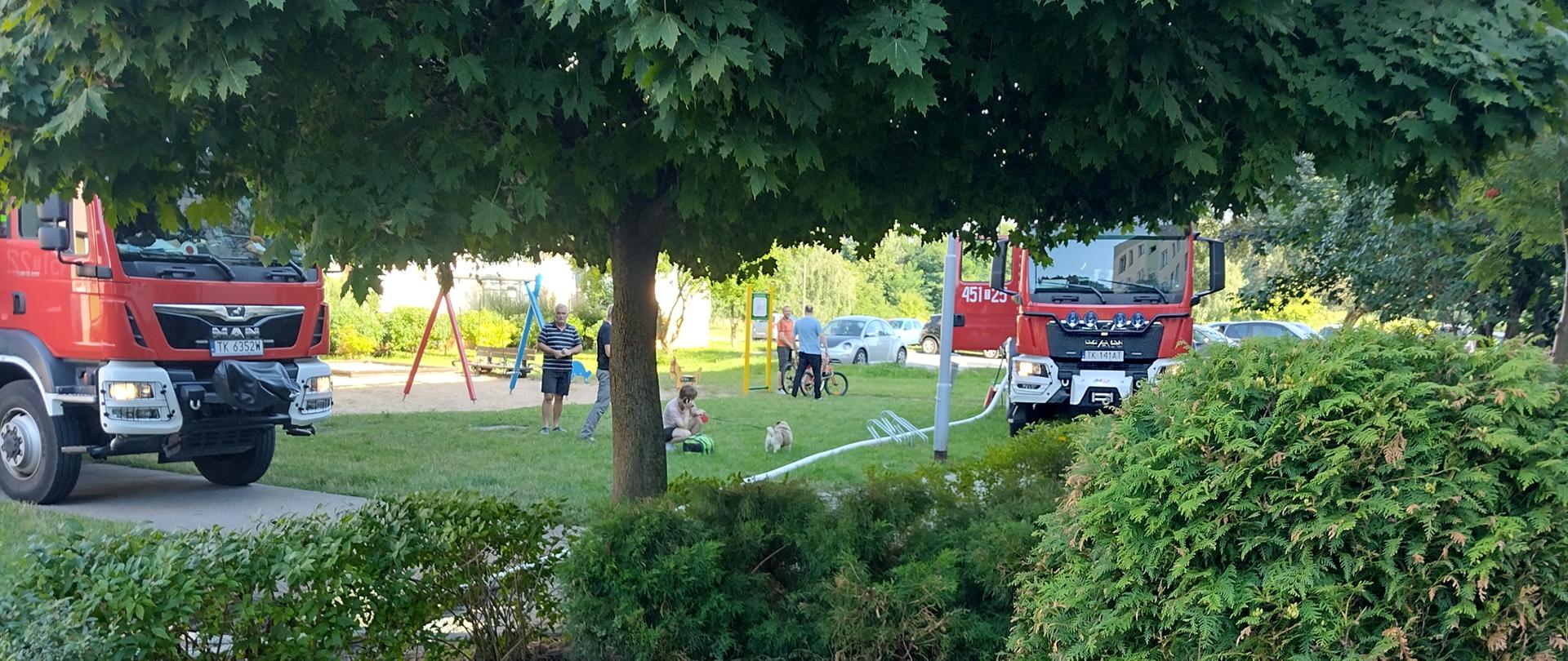 Na zdjęciu w porze dziennej widać przed wejściem do klatki schodowej do budynku w którym doszło do pożaru rozwinięta linię gaśniczą oraz dwa samochody strażackie. W tle plac zabaw przed blokiem