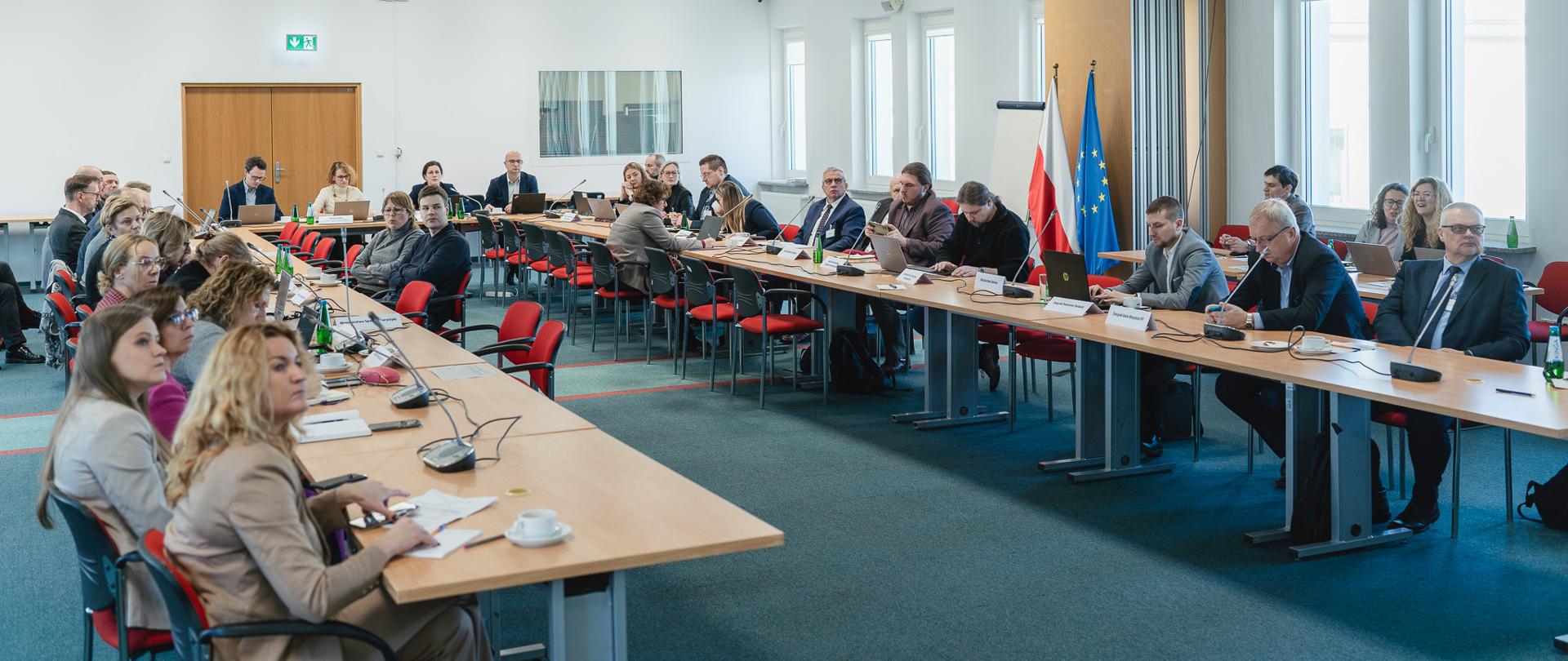 Grupa osób siedzi na sali wokół stołów ułożonych prostopadle do siebie.