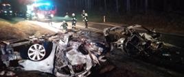 Na tle lasu widać pozostałości po samochodach osobowych które zapaliły się po ich zderzeniu. Auto znajdujące się po lewej stronie leży w na dachu poza jezdnią. Strażacy w tle zabezpieczają miejsce zdarzenia.