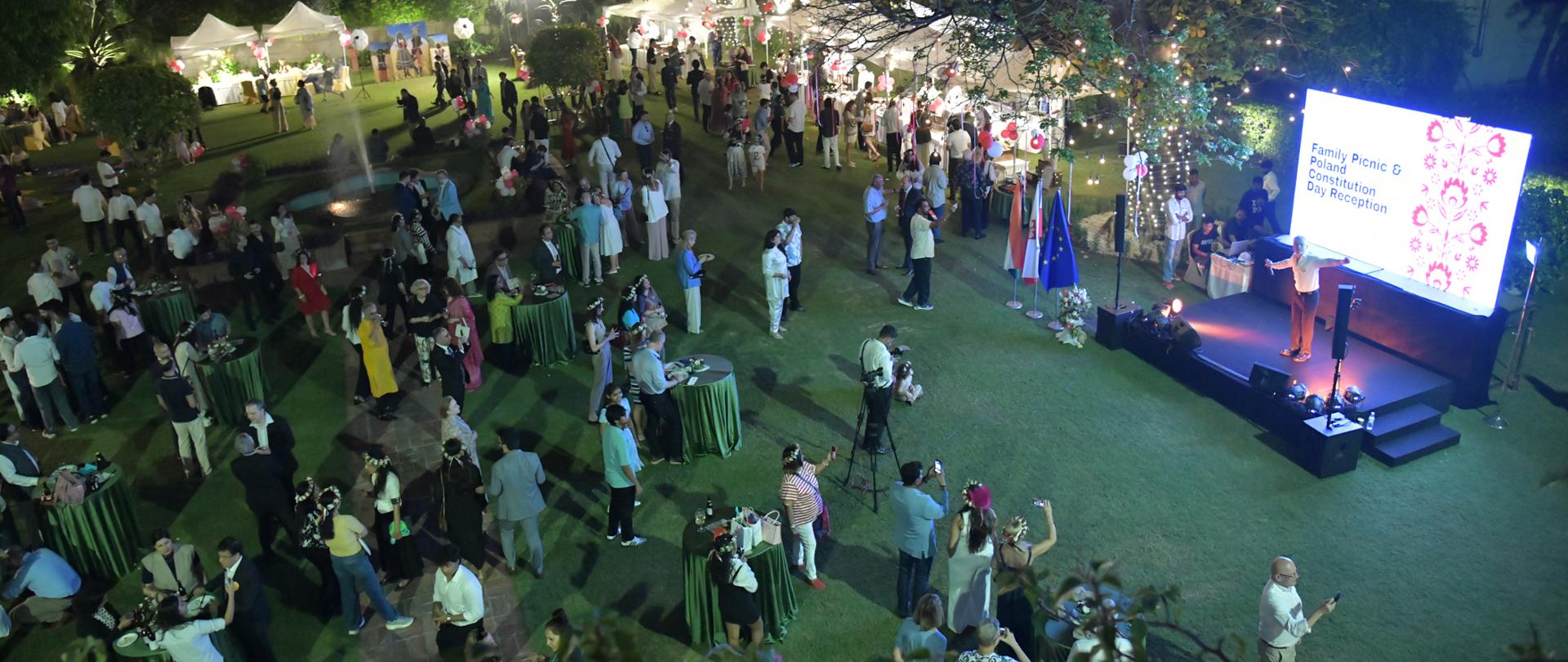 An evening birds-eye view of a lively outdoor reception for Poland’s Constitution Day. Guests gather around tables and tents, while artist Michał Rudaś stands on a lit stage. 