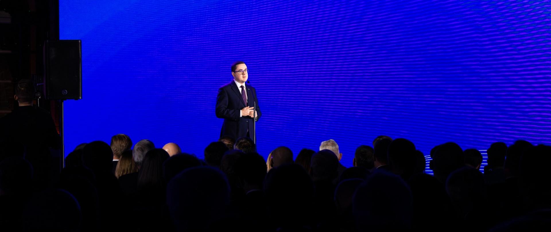 Minister Energii Miłosz Motyka przemawia na scenie przed dużą publicznością; za nim widać ogromny niebieski ekran LED i rząd reflektorów, a w tle ceglane ściany.