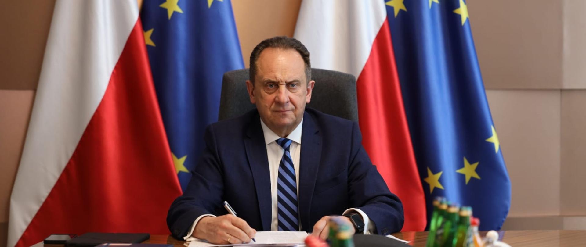 Minister Gut-Mostowy przy stole na tle flag PL i UE