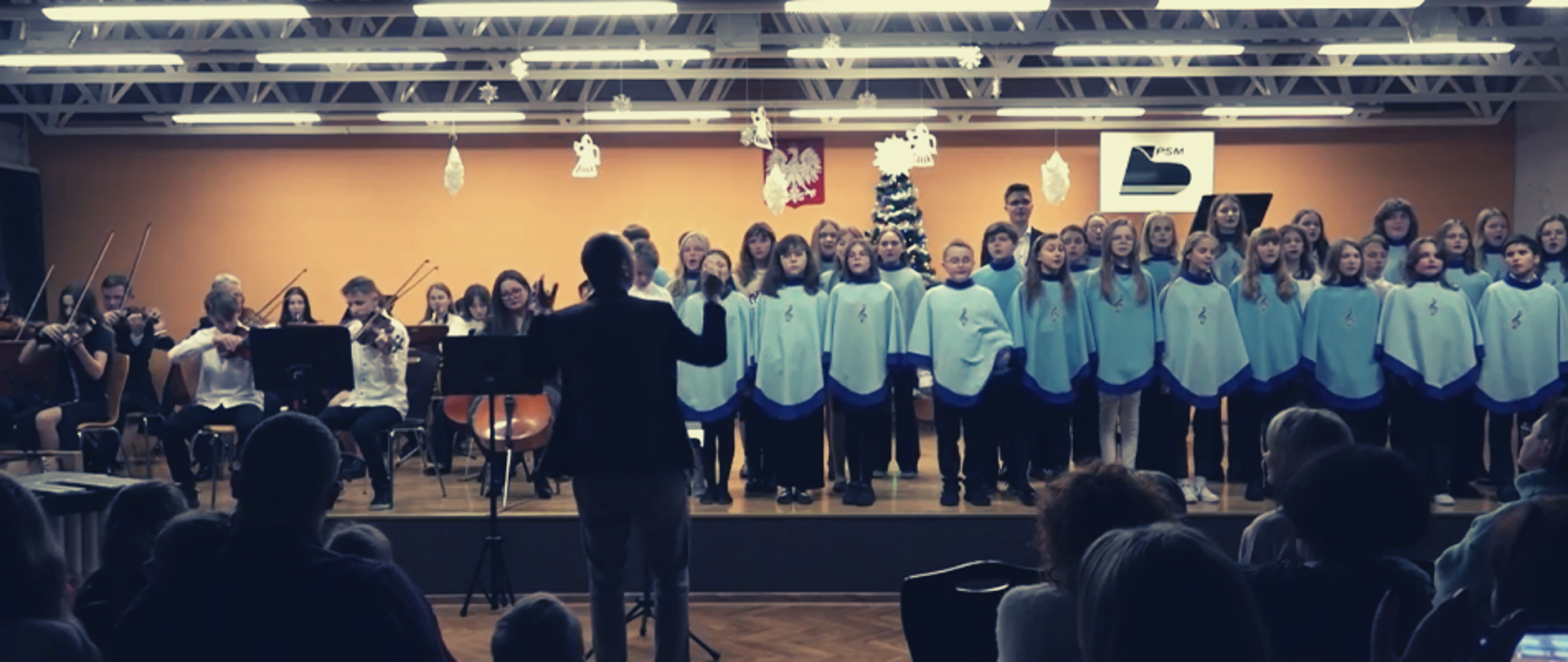 Zdjęcie przedstawia występ uczniów oraz nauczycieli podczas koncertu kolęd. Na scenie występują: chór szkolny oraz orkiestra stworzona na tą okoliczność. Całością dyryguje nauczyciel przedmiotów teoretycznych. Na zdjęciu widzimy również sylwetki przybyłych gości. 