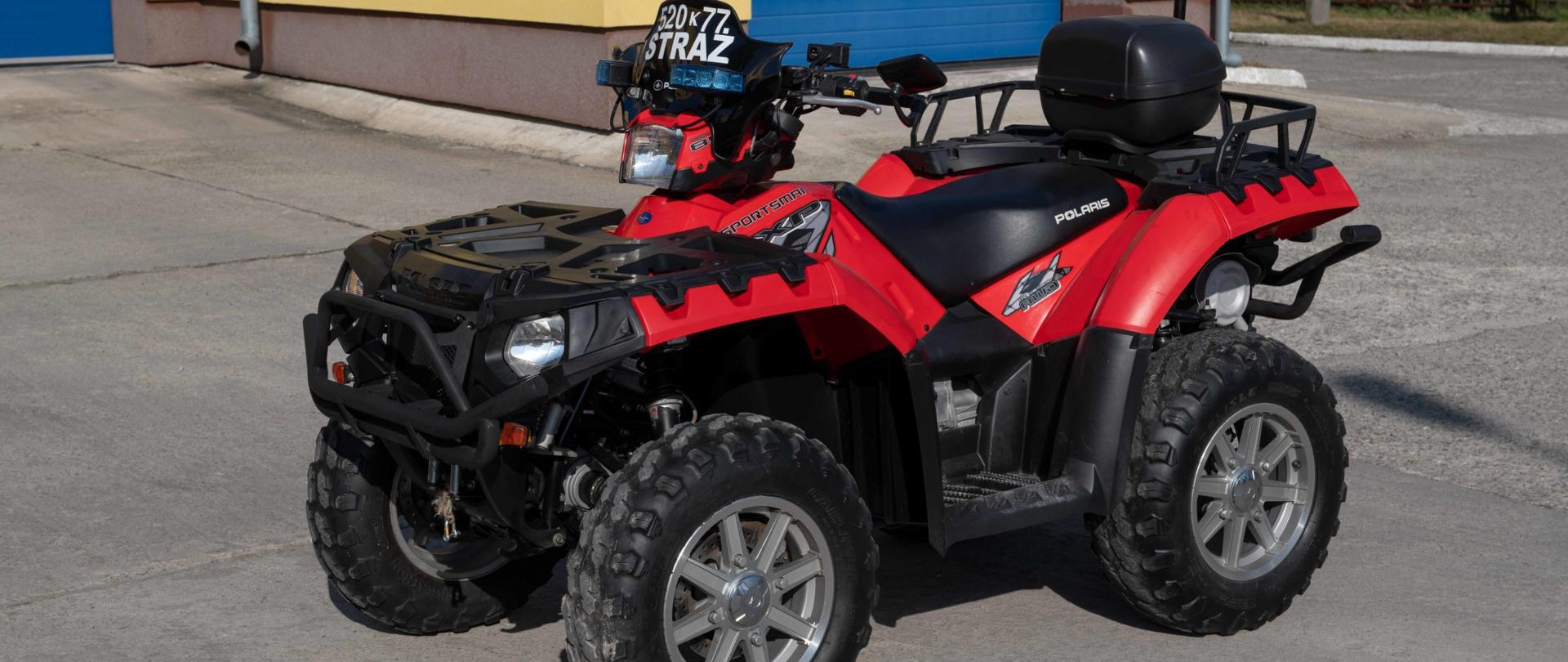 Dwuosiowy pojazd czerwono czarny marki polaris z terenowym ogumieniem bez osprzętu z kufrem za siedzeniem kierowcy. Na tle bram wjazdowych do garażu od strony placu wewnętrznego. 
