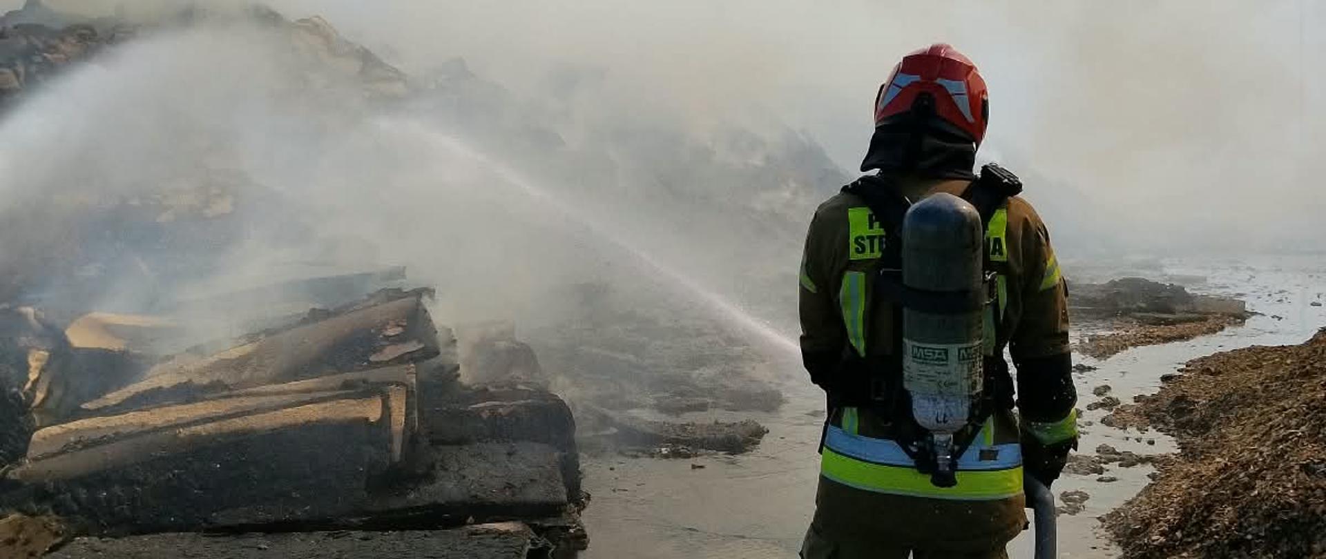 Na zdjęciu widać strażaka od tyłu, który trzyma wąż gaśniczy i kieruje strumień wody na płonącą pryzmę. Strażak jest ubrany w pełny strój ochronny i ma na plecach aparat oddechowy. Nad płonącym materiałem wznosi się gęsty dym.