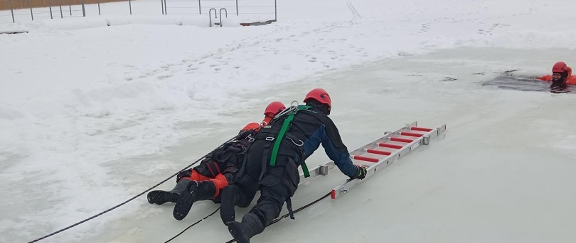 Zdjęcie obrazuje 2 ratowników przemieszczających się na czworakach na lodzie z drabiną w kierunku przerębla gdzie jest osoba w wodzie.