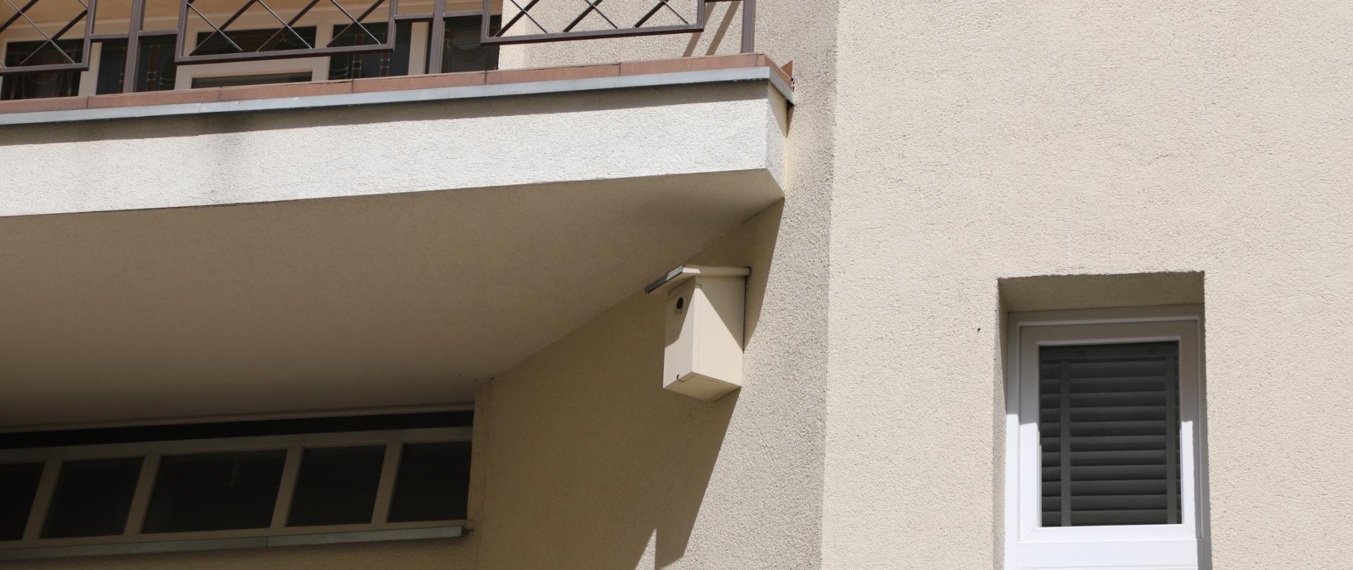 Skrzynka lęgowa typu A zamontowana na budynku i pomalowana w kolorze elewacji, tj. na beżowo. Na fotografii widać też fragmenty balkonu i okien.