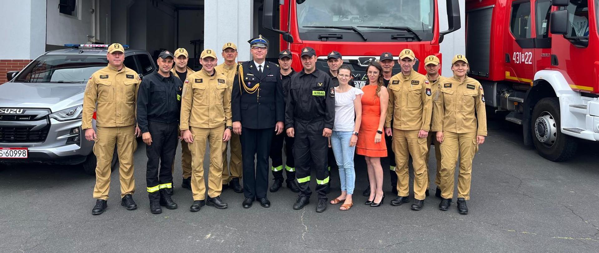 Pracownicy komendy stoją przed komendą. W tle stoją pojazdy pożarnicze.