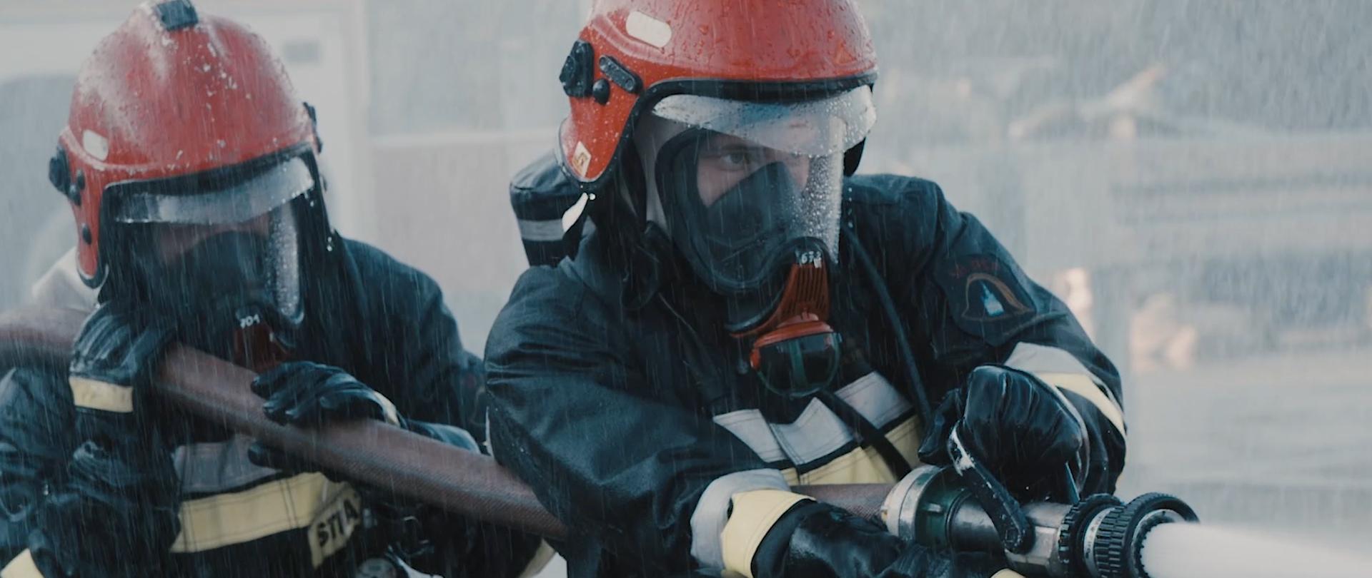 Kadr z filmu promującego Szkołę Aspirantów Państwowej Straży Pożarnej w Poznaniu. Na zdjęciu strażacy w ubraniu specjalnym i w sprzęcie ochrony dróg oddechowych gaszą pożar. Strażak po prawej stronie trzyma prądownicę i operuje prądem wodnym. Strażak po lewej stronie podtrzymuje wąż. Pada deszcz.