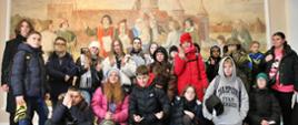Grupa kolorowo ubranej młodzieży stoi pod ścianą z freskiem przedstawiającym Mikołaja Kopernika i zamek.