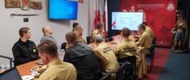 Zdjęcie przedstawia strażakom siedzących przy stole w sali konferencyjnej. Zastępca komendanta miejskiego Państwowej Straży Pożarnej omawia prezentację dotyczącą łączności.