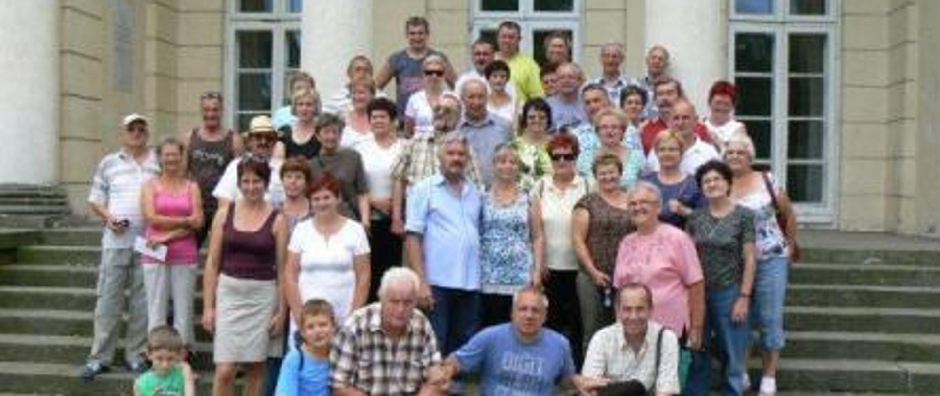 Na zdjęciu widzimy kobiety oraz mężczyzn pozujących w grupie do zdjęcia na schodach przed zabytkowym budynkiem koloru piaskowego. Dalej za grupą pozujących widzimy trzy kolumny budynku koloru białego, trzy okna balkonowe. 