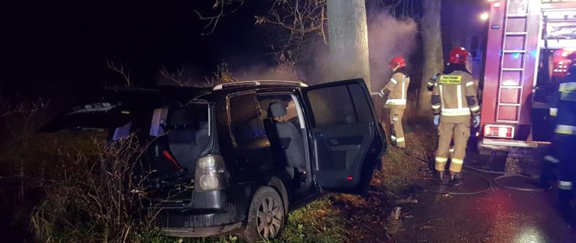 Widoczny od tyłu samochód osobowy który uderzył w drzewo. Auto koloru czarnego. Ma otwarte drzwi, z auta unosi się gęsty dym. Auto przodem uderzyło w drzewie. Obok stoi pojazd gaśniczy i strażacy