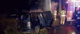 Widoczny od tyłu samochód osobowy który uderzył w drzewo. Auto koloru czarnego. Ma otwarte drzwi, z auta unosi się gęsty dym. Auto przodem uderzyło w drzewie. Obok stoi pojazd gaśniczy i strażacy