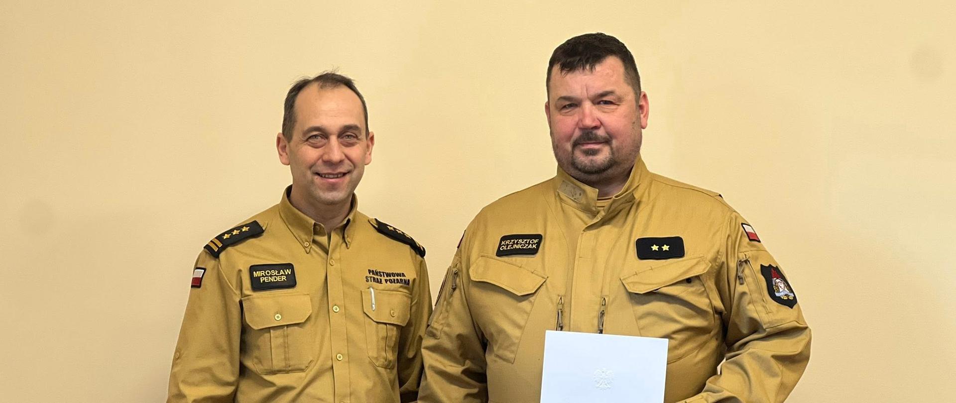 Dwóch strażaków w jasnych mundurach stoi obok siebie w pomieszczeniu. Jeden z nich trzyma dokument.