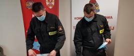Dwóch umundurowanych strażaków prezentujących ulotki z akcji szczepień przeciwko COVID-19