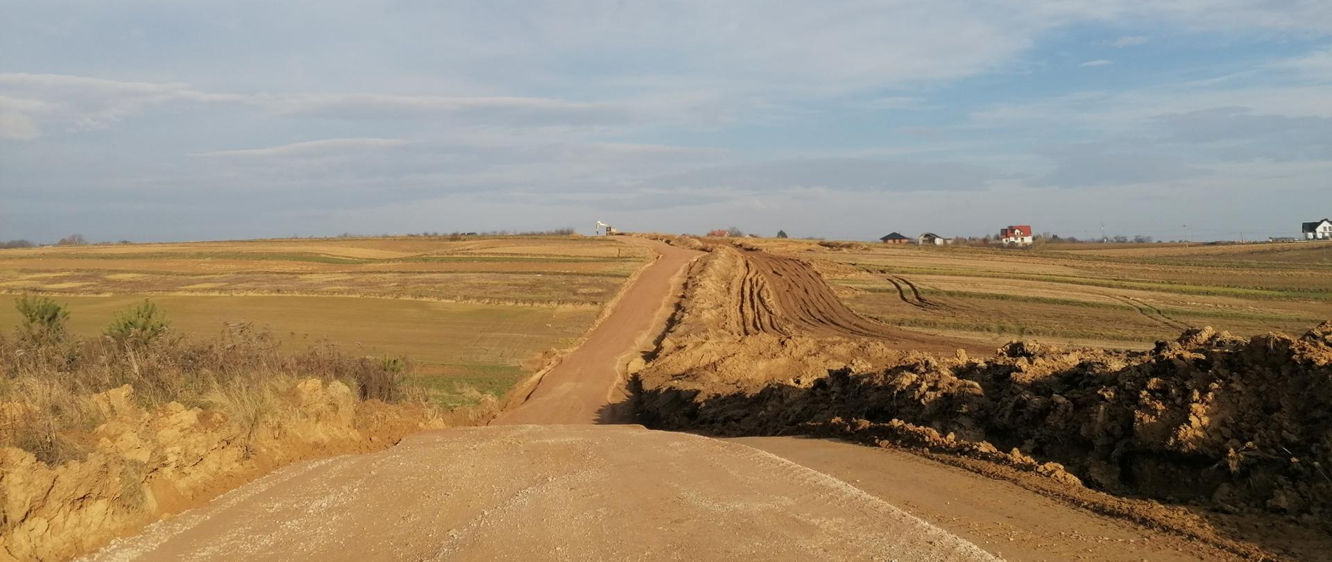 Zdjęcie budowanej drogi. Droga wznosi się na niewysokie wzniesienie pomiędzy polami uprawnymi. Na wzniesieniu, będącego linią horyzontu widać w tle maszynę budowlaną przy drodze oraz kilka budynków mieszkalnych. Bezśnieżna zima, wczesny ranek, słońce za plecami, niebo pokryte prześwitującymi szarymi chmurami.