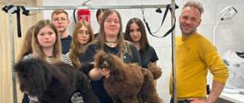 Zdjęcie przedstawia grupę siedmiu osób pozujących w salonie groomerskim (fryzjerskim dla zwierząt). W centrum uwagi znajdują się dwa profesjonalnie ostrzyżone psy rasy pudel, stojące na niebieskim stole groomerskim.Kompozycja i osobyPo prawej stronie stoi uśmiechnięty mężczyzna w wieku około 35-40 lat. Ma krótkie, jasne, postrzępione włosy, kilkudniowy zarost i żółty, dopasowany sweter oraz jeansy. Trzyma dłoń na biodrze.W centralnej części i po lewej stronie stoi grupa sześciu nastolatków (pięć dziewcząt i jeden chłopiec w okularach). Wszyscy patrzą w stronę obiektywu, mają neutralne lub lekko uśmiechnięte miny i są ubrani w ciemną odzież (głównie czarne koszulki).Jedna z dziewcząt w centrum trzyma oburącz mniejszego, brązowego psa, stabilizując go na stole.Psy na pierwszym planiePo lewej stronie stoi większy pudel o bardzo ciemnym, niemal czarnym futrze. Ma charakterystyczną fryzurę z dużą, puszystą "czapą" na głowie i obfitymi, szerokimi nogawkami z futra na łapach. Pies jest zwrócony bokiem w lewą stronę.Po prawej stronie stoi mniejszy pudel o jasnobrązowej (czekoladowej) sierści. Jest zwrócony w prawą stronę, ale odwraca głowę prosto w stronę aparatu. Jego sierść również jest precyzyjnie przystrzyżona, odsłaniając szczupłą sylwetkę i zostawiając puszyste pompony na łapach i głowie.Tło i otoczenieAkcja toczy się w jasnym pomieszczeniu o ścianach wyłożonych dużymi, białymi lub jasnoszarymi kafelkami z połyskiem.Niebieski stół, na którym stoją psy, ma metalową konstrukcję (szubienicę groomerską) z czarnymi smyczami stanowiskowymi zwisającymi nad zwierzętami.W tle na ścianie, dokładnie nad głowami nastolatków, wisi czerwona gaśnica oraz czerwony znak przeciwpożarowy.Po prawej stronie widoczna jest biała, specjalistyczna lampa biurkowa oraz fragment stanowiska pracy z białym blatem.Jeśli chcesz poznać więcej szczegółów, chętnie opiszę fryzury psów, dokładny ubiór konkretnych osób lub elementy wyposażenia salonu.