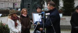 Na dworze, minister Czarnek w czarnej kurtce trzyma oprawiony w ramki dokument, przed nim stoją trzy kobiety, w tle czarny pomnik na placu.