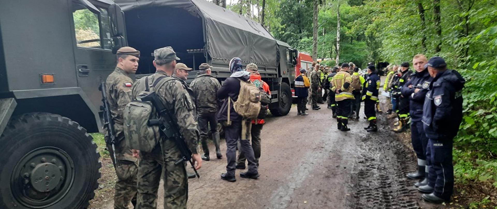 Żołnierze WOT strażacy PSP i OSP i policjanci stoją w grupach w tle pojazdy wojska i straży.
