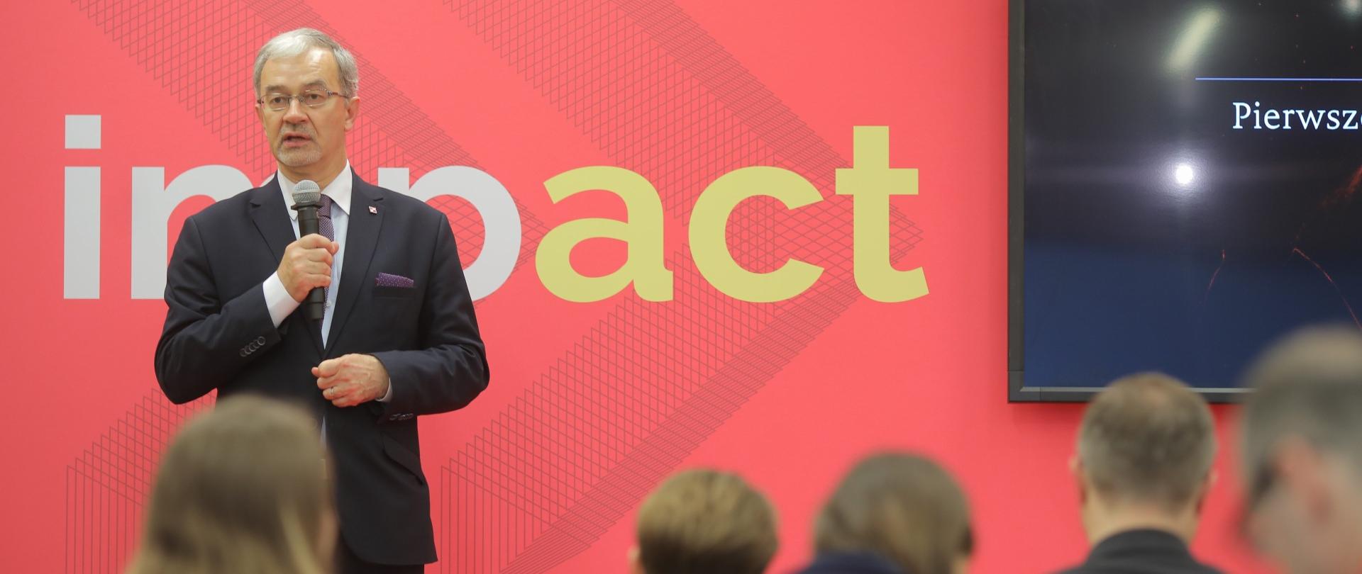 Minister Jerzy Kwieciński stoi z mikrofonem w ręku na tle ekranu z napisem "impact". Przed nim siędza osoby, uczestnicy konferencji.