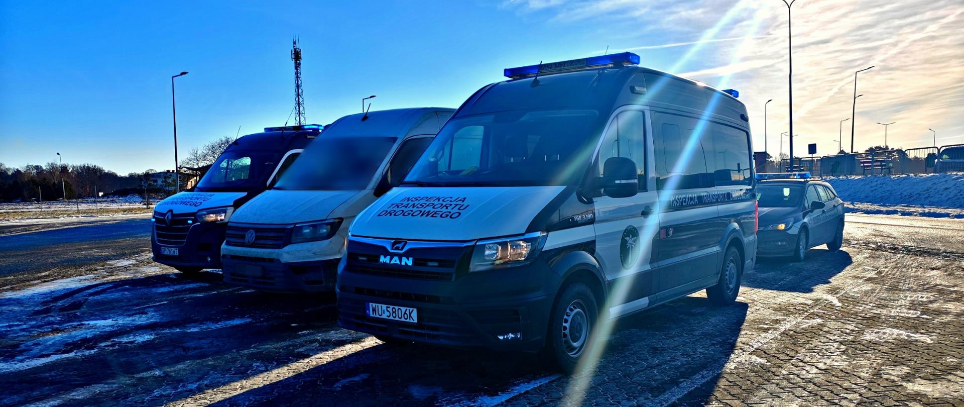 Radiowozy Inspekcji Transportu Drogowego oraz pojazd Krajowej Administracji Skarbowej stojące na parkingu zimą, z niebieskimi światłami ostrzegawczymi, na tle jasnego nieba i niskiego słońca.