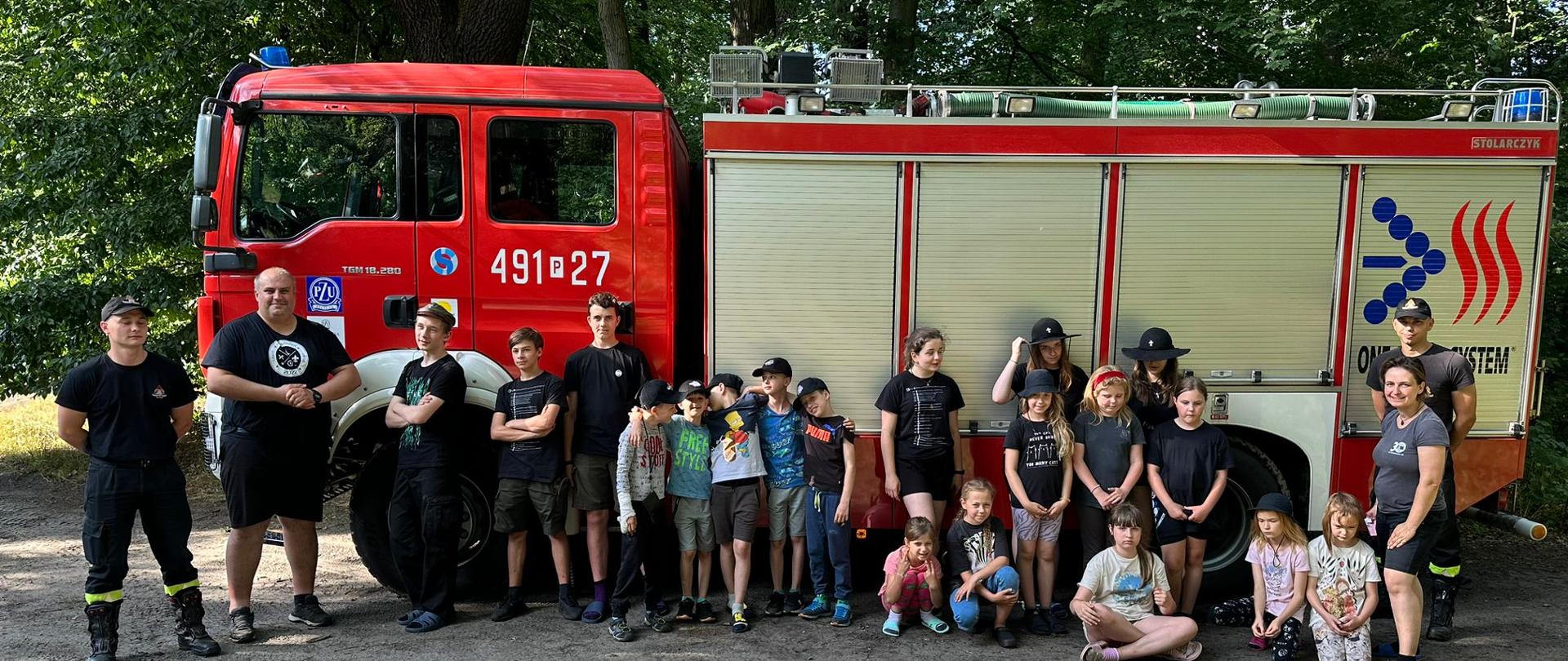 Zdjęcie grupowe przedstawiające uczestników obozu harcerskiego oraz strażaków uczestniczących w rozpoznaniu obozowiska. Zdjęcie wykonane w kompleksie leśnym na tle pojazdu pożarniczego. 