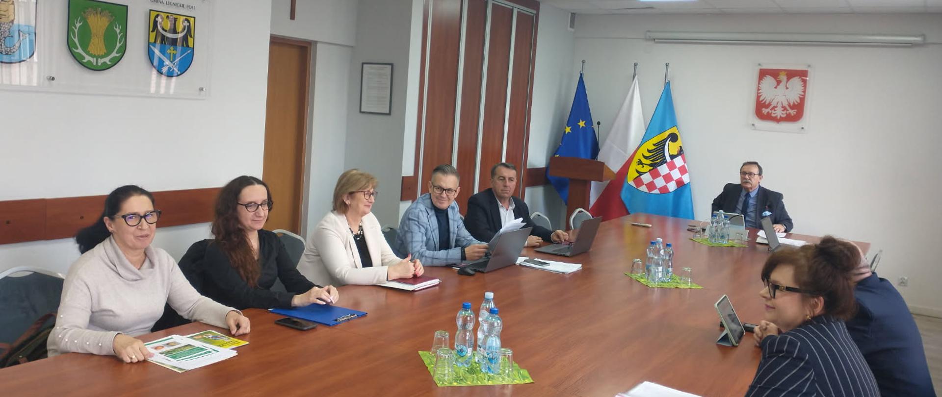 Spotkanie w sali konferencyjnej, kilka osób siedzi przy stole z dokumentami i laptopami; w tle widoczne flagi Polski i Unii Europejskiej oraz herby na ścianie.