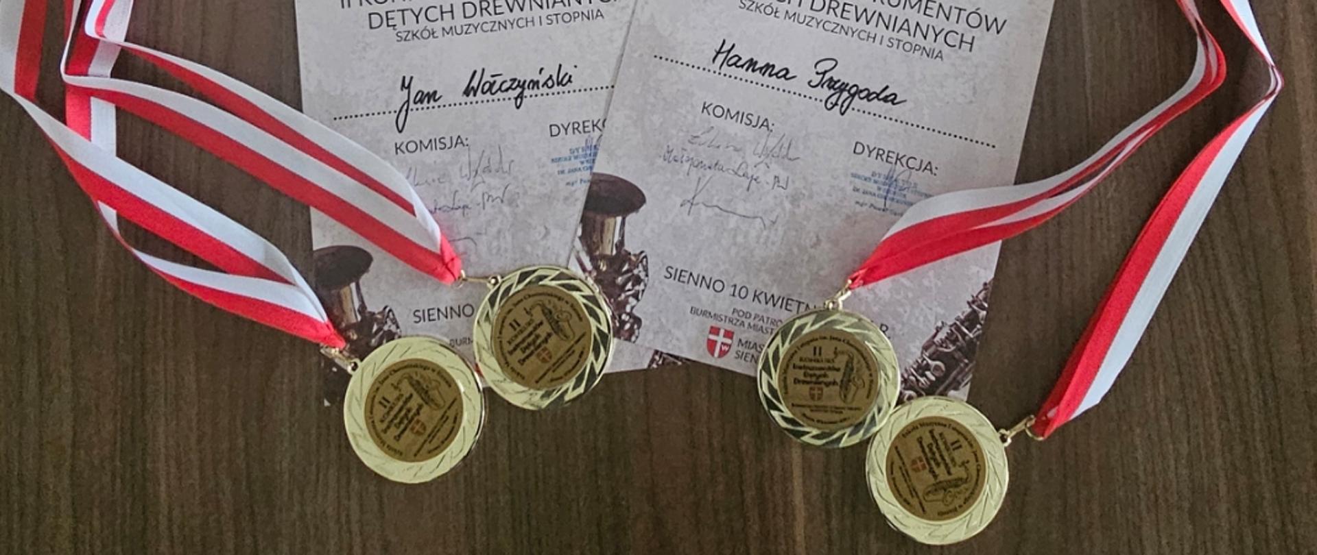 Pięć dyplomów ułożonych na drewnianym blacie, przyznanych za udział w II Konkursie Instrumentów Dętych Drewnianych Szkoły Muzycznej I Stopnia im. Jana Chorosińskiego w Sienne. Każdy dyplom zawiera imię i nazwisko laureata oraz podpisy komisji i dyrekcji. Obok dyplomów leżą trzy złote medale zawieszone na czerwono-białych wstążkach, symbolizujących osiągnięcia i wyróżnienia.