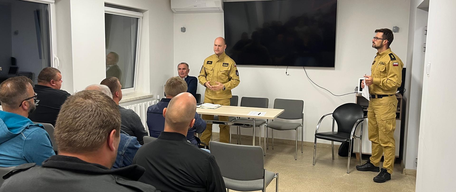 Na sali siedzą mężczyźni na krzesłach, przed nimi stoi dwóch strażaków w umundurowaniu w kolorze piaskowym, obok jednego z nich siedzi pan w marynarce.