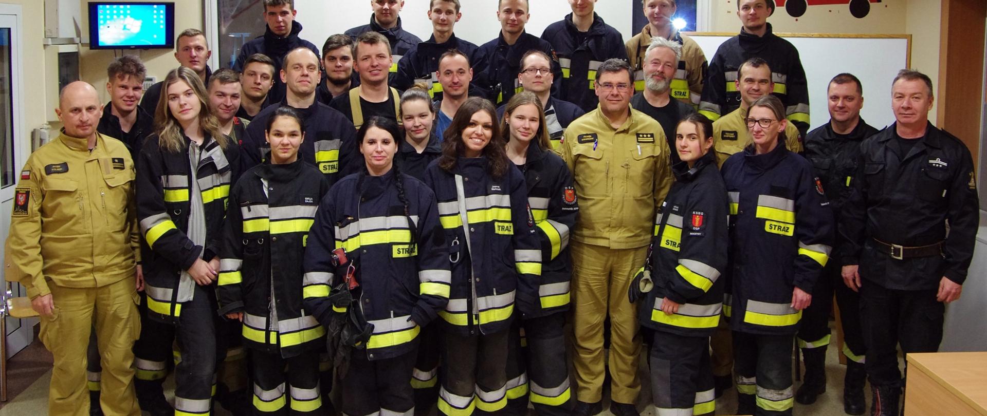 Widok ogólny z przodu – zdjęcie zbiorowe. Absolwenci i kadra PSP stoją na sali szkoleniowej.