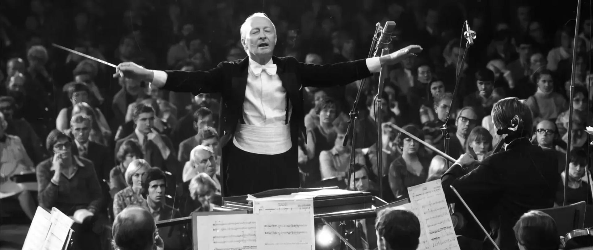 Zdjęcie przedstawiające na głównym planie Witolda Lutosławskiego jako dyrygenta orkiestry. Przed nim znajdują się instrumentaliści, z kolei za nim - widownia. Zdjęcie wykonano w trakcie koncertu.