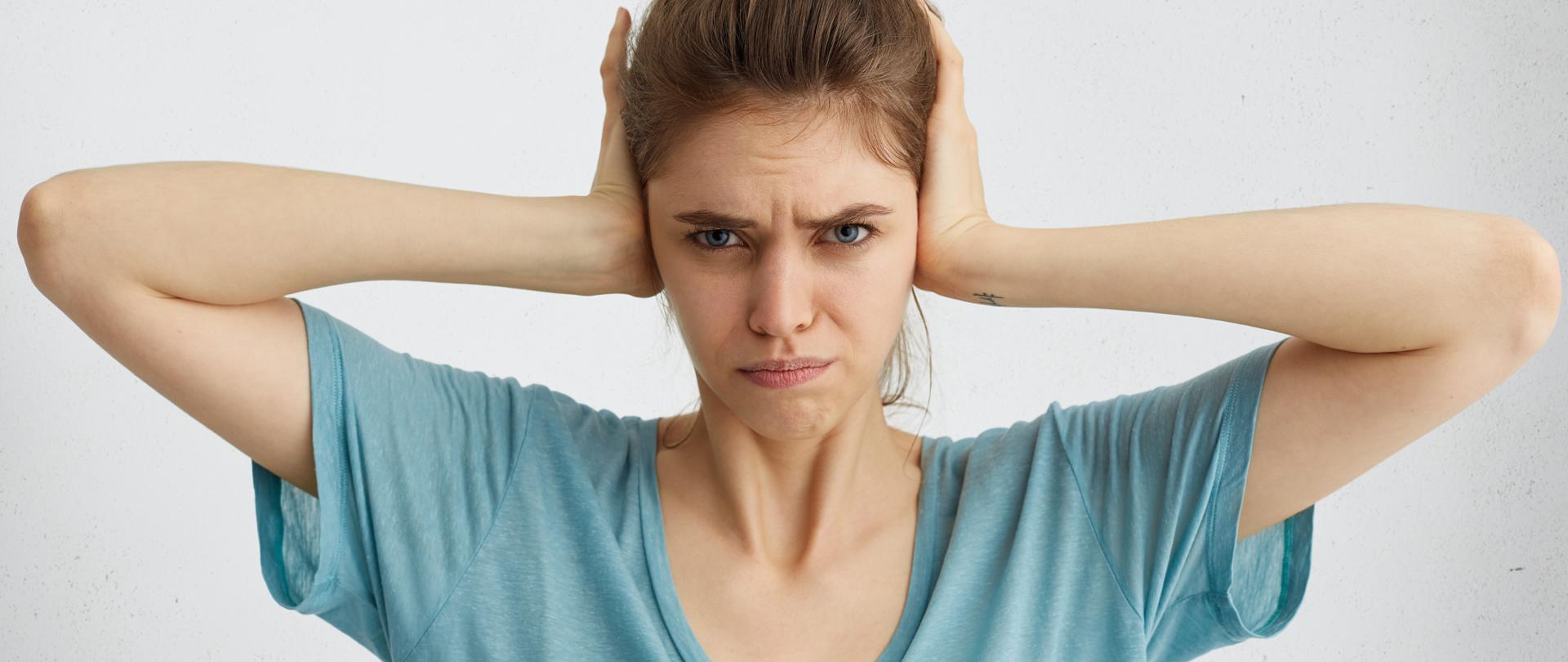 Zdjęcie przedstawia kobietę, która przebywa w hałasie. Zatyka uszy, aby nie słyszeć dźwięków.