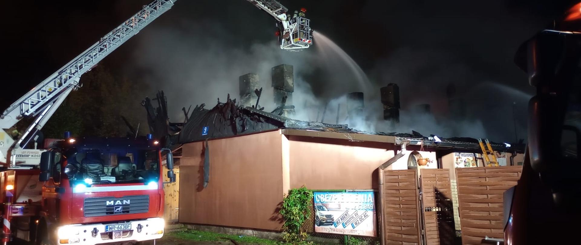 Zdjęcie wykonane w porze nocnej. Na zdjęciu widać pojazd pożarniczy podnośnik hydrauliczny. Pojazd w kolorze czerwonym. Pojazd posiada rozłożone ramię – wysięgnik zakończony koszem. W koszu znajdują się strażacy. Strażacy z kosza leją wodę na spalony i zawalony już dach budynku w którym wybuch pożar. 