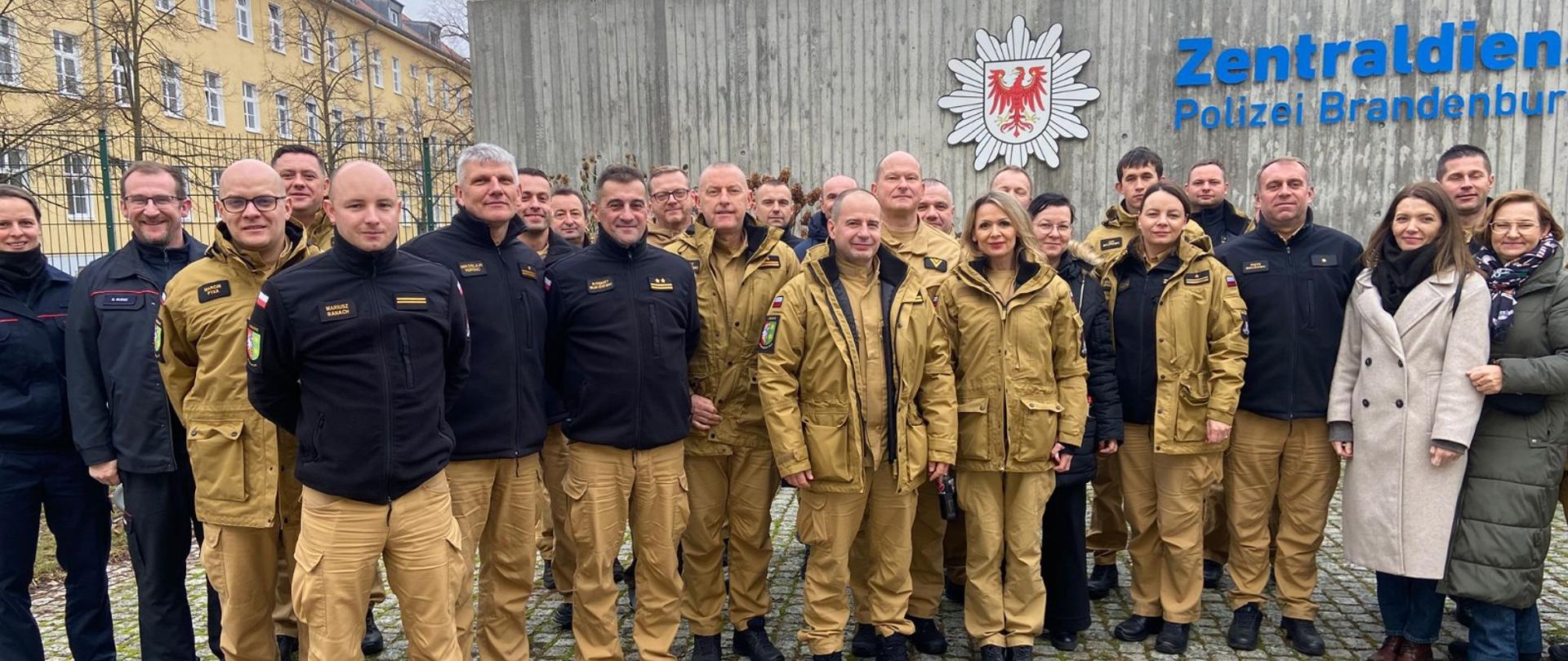 Uczestnicy wizyty studyjnej z Polski i Niemiec stoją wspólnie przed budynkiem Zentradienst Polizei Brandenburg. Widoczne są mundury PSP oraz mundury służb niemieckich.