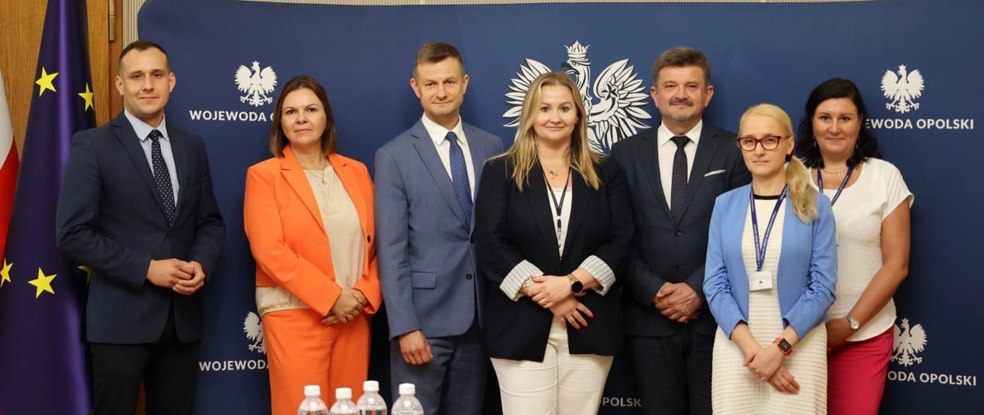 Uczestnicy spotkania stoją przed ścianką z napisem "Wojewoda Opolski" w Sali Złotej. Pozują do wspólnego zdjęcia po spotkaniu. 