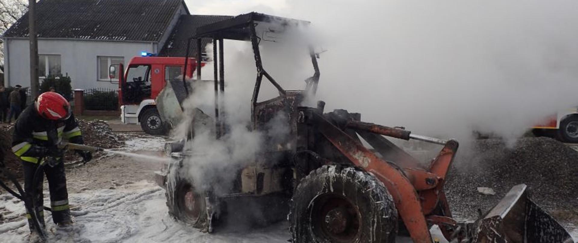 Pożar ładowarki w miejscowości Tarczek. Na pierwszym planie widoczna dymiąca się ładowarka oraz gaszący ją funkcjonariusz PSP. W tle widoczny samochód bojowy PSP.