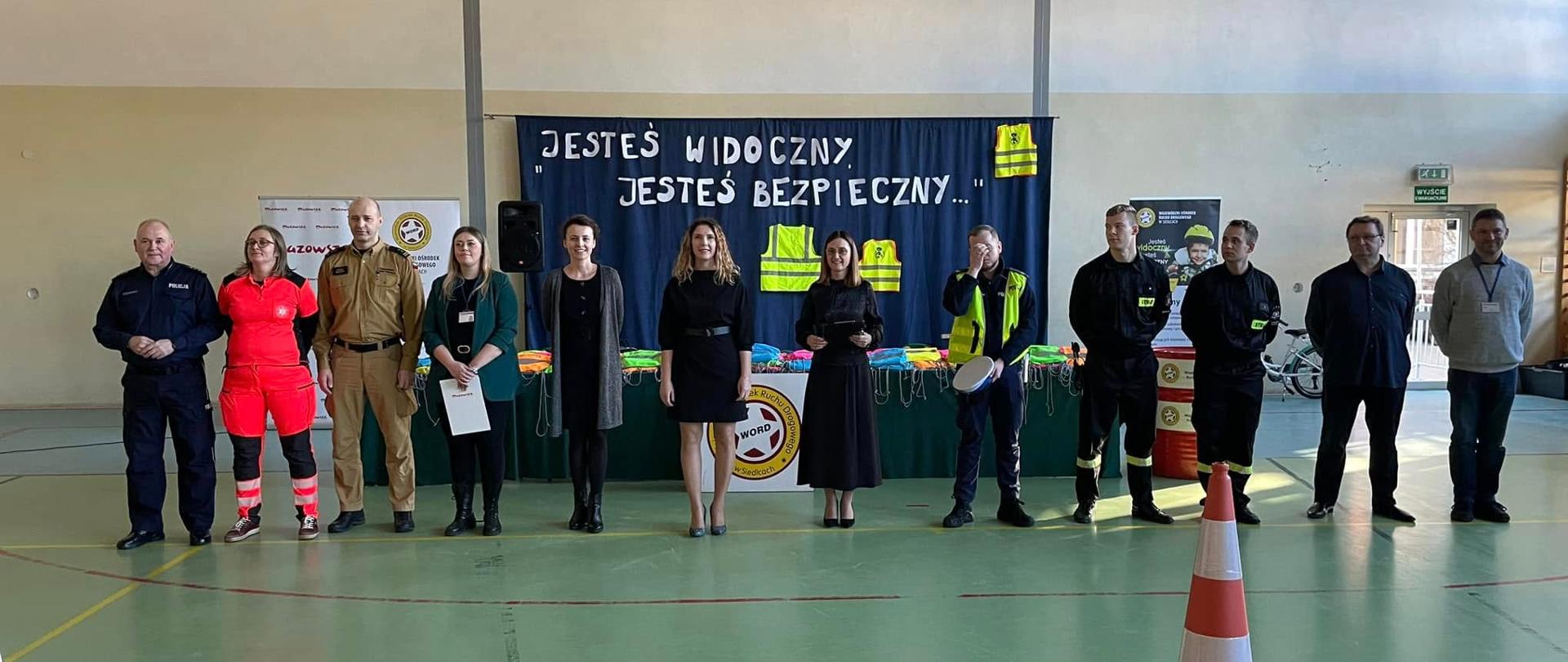 Z przyjemnością opiszę to zdjęcie.
**Zdjęcie c9cf6780-6de7-4202-93ed-47daf0fb09df.jpg:**
Zdjęcie przedstawia grupę jedenastu dorosłych osób, stojących w szeregu w sali gimnastycznej, prawdopodobnie podczas uroczystości lub podsumowania kampanii społecznej. W tle wisi duży, niebieski baner z białym napisem "JESTEŚ WIDOCZNY JESTEŚ BEZPIECZNY..." oraz symbolami odblaskowymi, co wskazuje, że wydarzenie dotyczy bezpieczeństwa i promowania noszenia odblasków.
Od lewej strony, w rzędzie stoją:
* Mężczyzna w ciemnym mundurze policyjnym.
* Kobieta w czerwonym kombinezonie ratowniczym.
* Mężczyzna w jasnobeżowym mundurze strażackim.
* Kobieta w ciemnym garniturze, trzymająca w ręku certyfikat lub dokument.
* Kobieta w czarnej sukience.
* Kobieta w czarnej sukience, również trzymająca dokument.
* Mężczyzna w żółtej kamizelce odblaskowej, trzymający w ręku białe przedmioty, prawdopodobnie kaski.
* Dwóch mężczyzn w ciemnych mundurach strażackich.
* Mężczyzna w granatowej bluzie.
* Mężczyzna w szarym swetrze i koszuli.
Na pierwszym planie, po prawej stronie, widać stożek drogowy, co dodatkowo podkreśla temat bezpieczeństwa drogowego. Cała scena ma charakter oficjalny i reprezentacyjny, związany z działaniami prewencyjnymi.