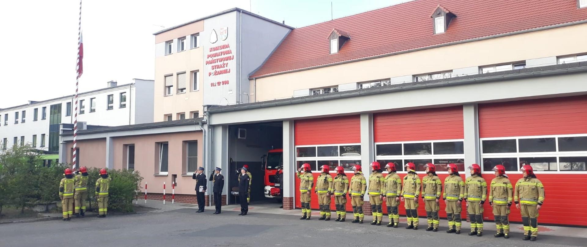 STRAŻACY ŚWIĘTUJĄ DZIEŃ FLAGI RZECZYPOSPOLITEJ POLSKIEJ. Na zdjęciu widać strażaków podczas uroczystego apelu z okazji Dnia Flagi, strażacy stoją w rzędzie, za nimi budynek strażnicy, flaga Polski uroczyście wciągana na maszt.