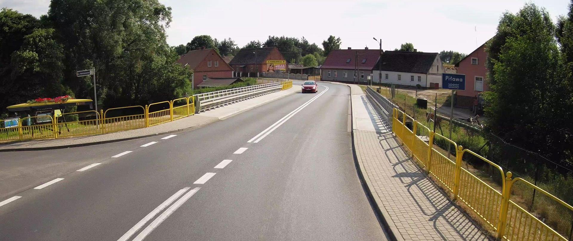 Droga w miejscowości z widocznym na pierwszym planie mostem przez rzekę.
