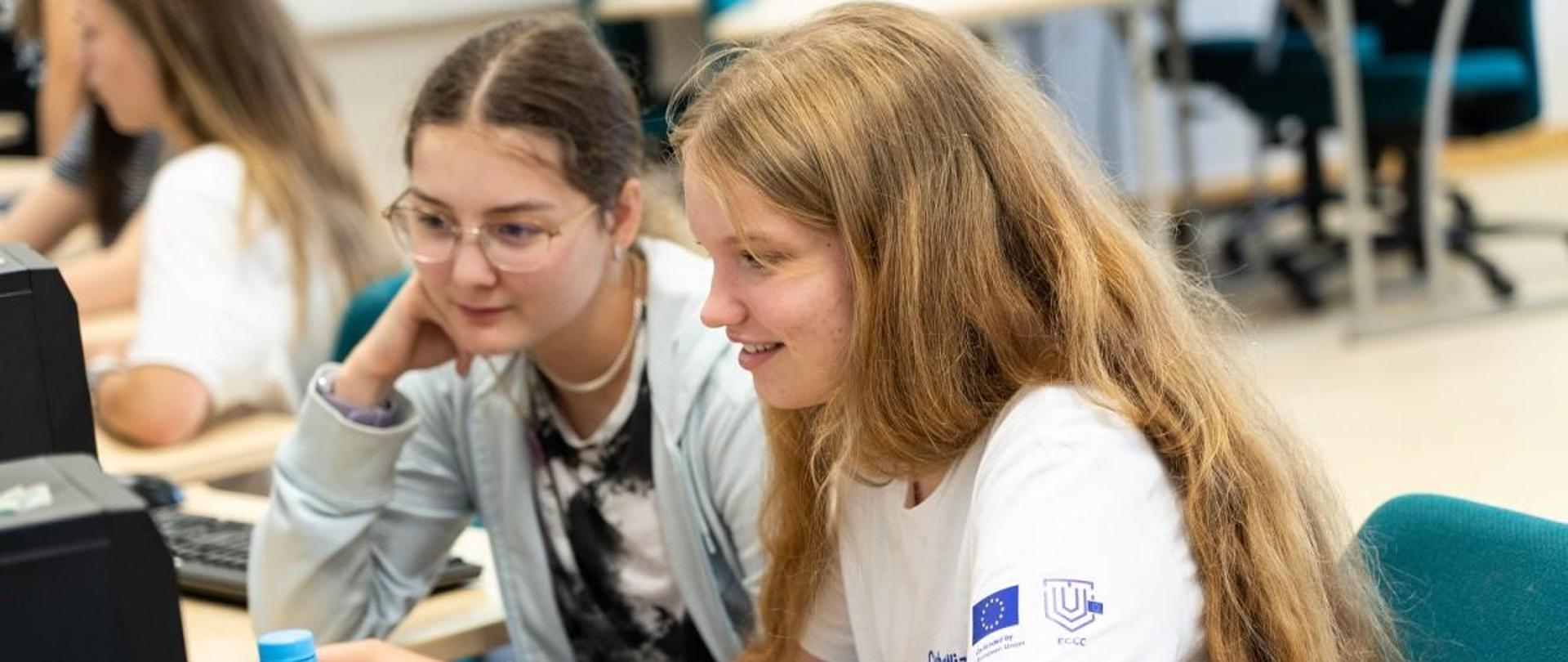 Dwie dziewczyny siedzą przy komputerze i patrzą w monitor, jedna z nich trzyma mysz w dłoni