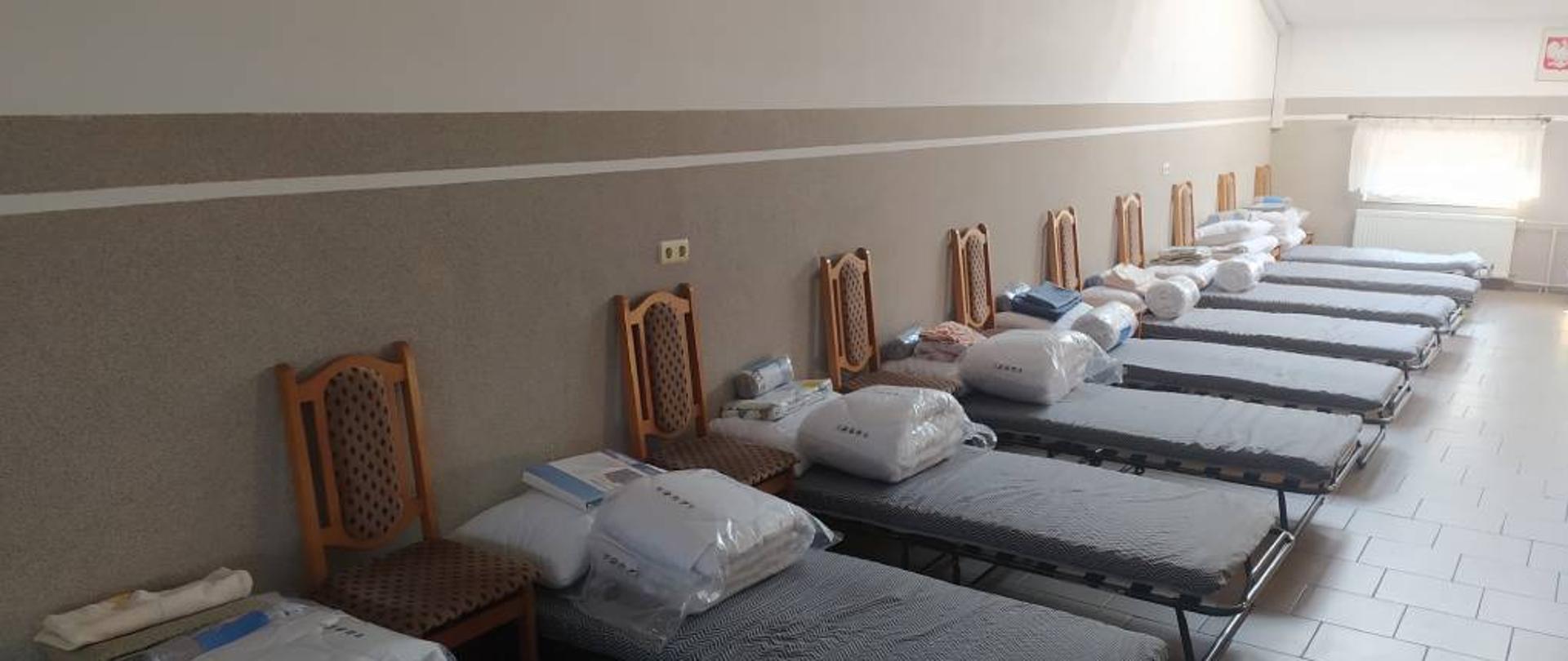 Zdjęcie przedstawia pomieszczenie przygotowane na przyjęcie uchodźców z Ukrainy. W znajdującym się na poddaszu budynku Ochotniczej Straży Pożarnej Kołakach Kościelnych widać ustawione przy jednej ze ścian łóżka polowe oraz krzesła. Na każdym z łóżek znajduje się komplet pościeli oraz ręcznik. Zdjęcie wykonano na poddaszu budynku Ochotniczej Straży Pożarnej Kołakach Kościelnych w słoneczny dzień.