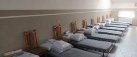 Zdjęcie przedstawia pomieszczenie przygotowane na przyjęcie uchodźców z Ukrainy. W znajdującym się na poddaszu budynku Ochotniczej Straży Pożarnej Kołakach Kościelnych widać ustawione przy jednej ze ścian łóżka polowe oraz krzesła. Na każdym z łóżek znajduje się komplet pościeli oraz ręcznik. Zdjęcie wykonano na poddaszu budynku Ochotniczej Straży Pożarnej Kołakach Kościelnych w słoneczny dzień.