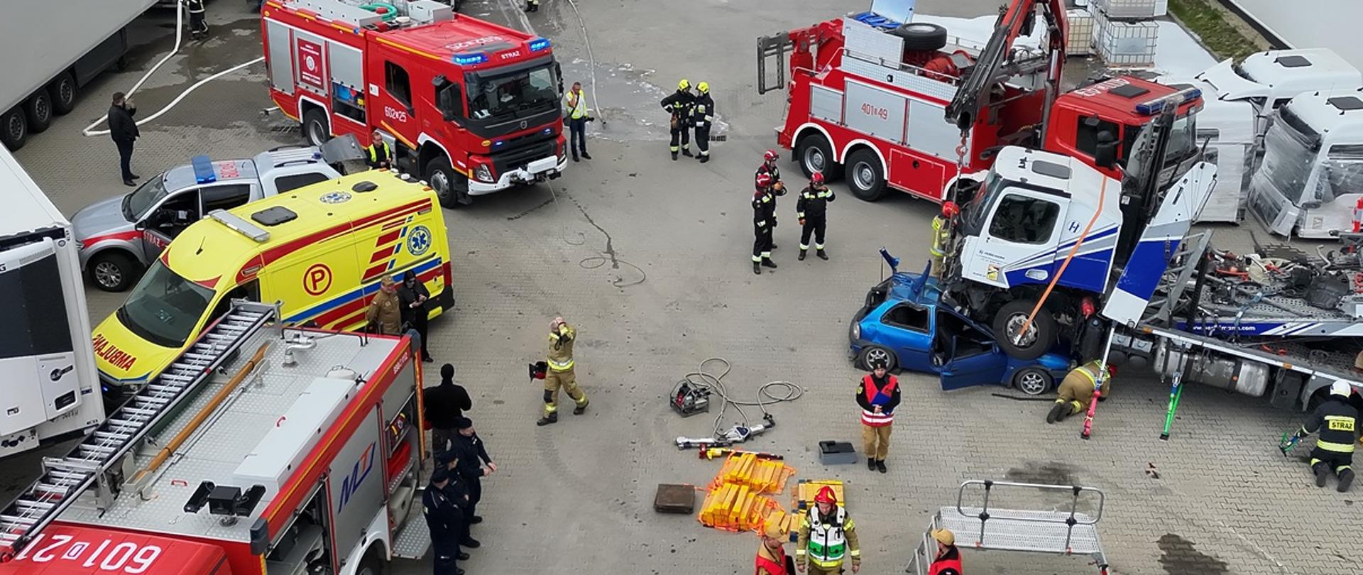 Dzień. Widok na plac manewrowy przy hali magazynowej. Na placu trwają ćwiczenia strażackie - zderzenie pojazdu osobowego w kolorze niebieskim z samochodem ciężarowym. Obok stoją strażacy oraz kilka pojazdów ratowniczo-gaśniczych i karetka.