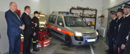 Przekazanie samochodu w OSP Jutrosin. Garaż w remizie OSP Jutrosin. Kierowca OSP uruchamia silnik przekazanego samochodu oraz sygnały dźwiękowe i świetle - na potwierdzenie, że wóz jest w pełni sprawny. Z lewej strony goście spotkania, a z prawej druhowie OSP.