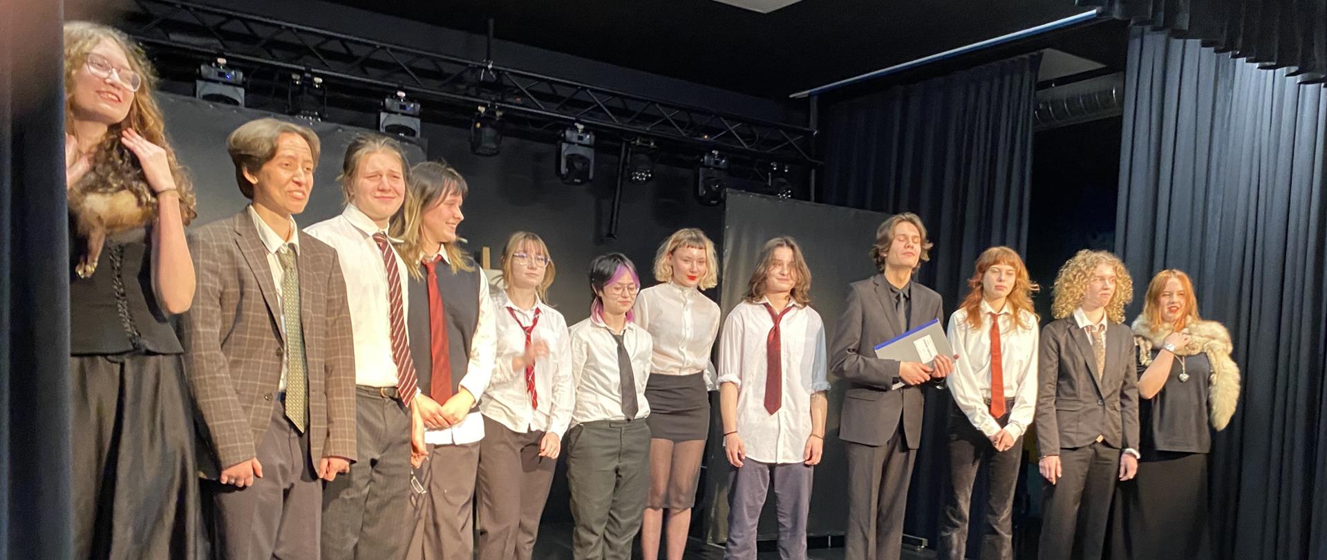 Na scenie stoi dwunastu młodych ludzi ubranych w koszule, garnitury, krawaty, a po bokach dwie dziewczyny w etolach z lisa