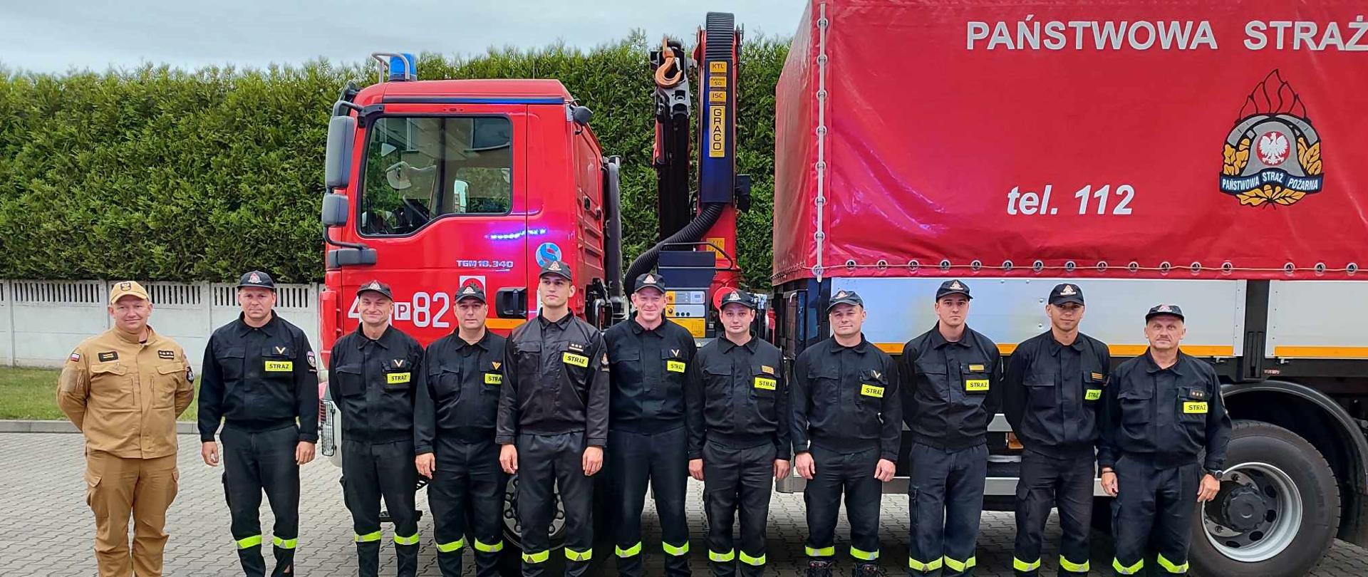 Zdjęcie przedstawia strażaków w umundurowaniu koszarowym na tle samochodu pożarniczego, który brał udział w misji ratowniczej w Grecji.